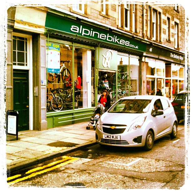 Silver car parked outside an Alpine Bikes storefront, with bicycles in the window and a person pushing a stroller on the sidewalk.
