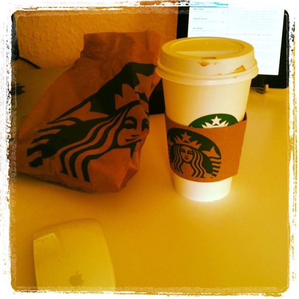 Starbucks paper bag and lidded coffee cup on a desk, with a computer screen in the background and a white mouse in the foreground.