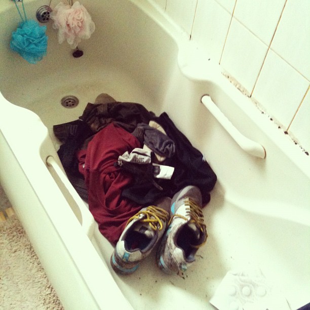 Pile of clothes and a pair of sneakers in a bathtub with two bath poufs hanging near the faucet.
