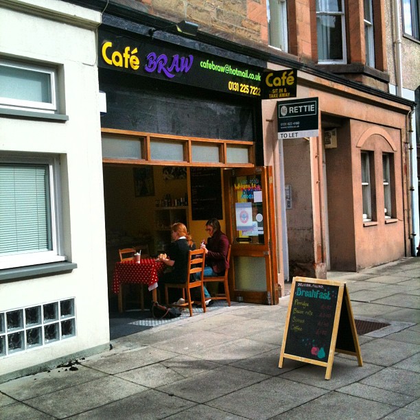 Two people sit at a small table inside an open-front cafe, with a breakfast chalkboard sign on the sidewalk.