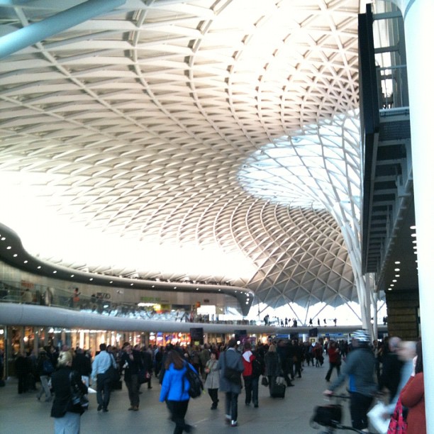 Crowded station concourse with people walking beneath a sweeping lattice roof and large glass opening.