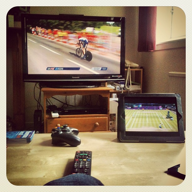 Living room with a TV showing a cyclist race, a tablet showing tennis, and a coffee table with a remote and game controller.