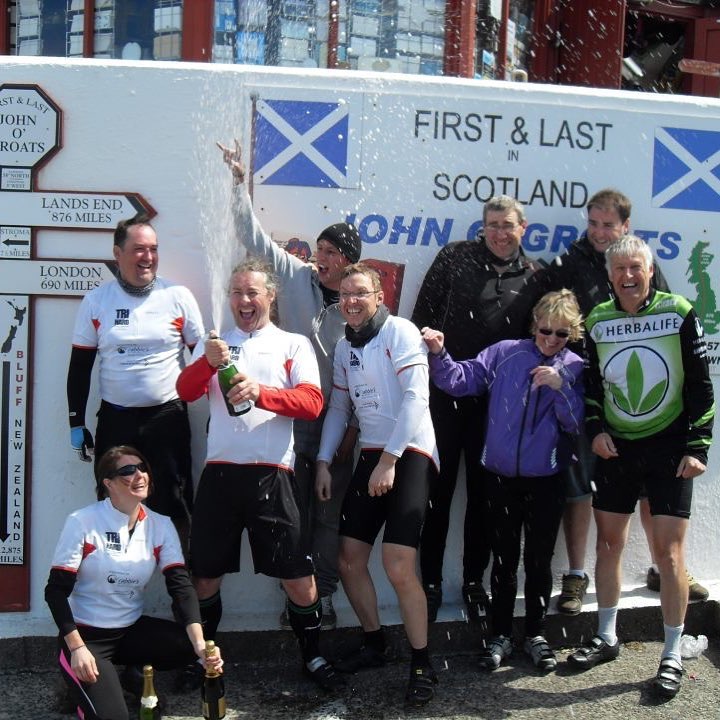Group of cyclists celebrating with champagne in front of a sign reading "First & Last in Scotland John O'Groats" with distance markers.