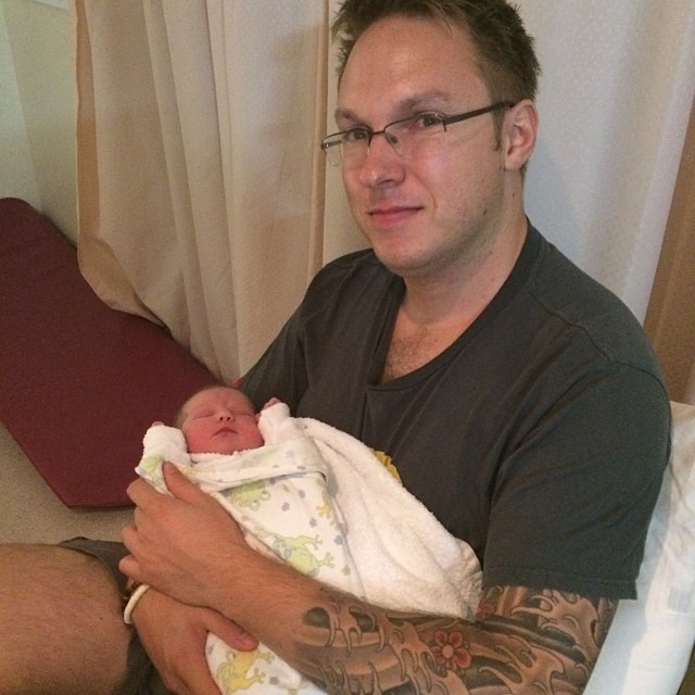 Man with glasses and tattooed arms holding a swaddled newborn wrapped in a white blanket.