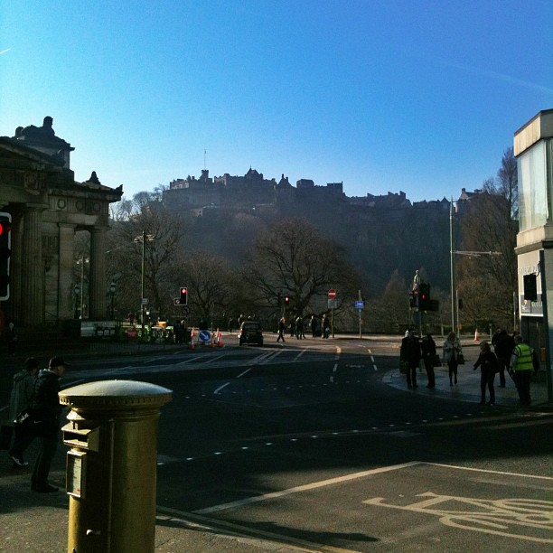 City street intersection with pedestrians and traffic lights, with a hilltop skyline of historic buildings silhouetted against a bright blue sky.