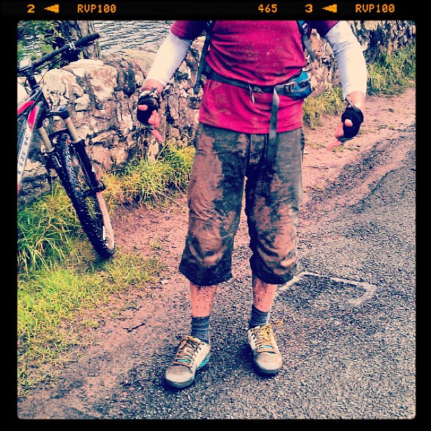 Person in muddy cropped pants, gloves, and sneakers standing on a wet road beside a bicycle and stone wall.