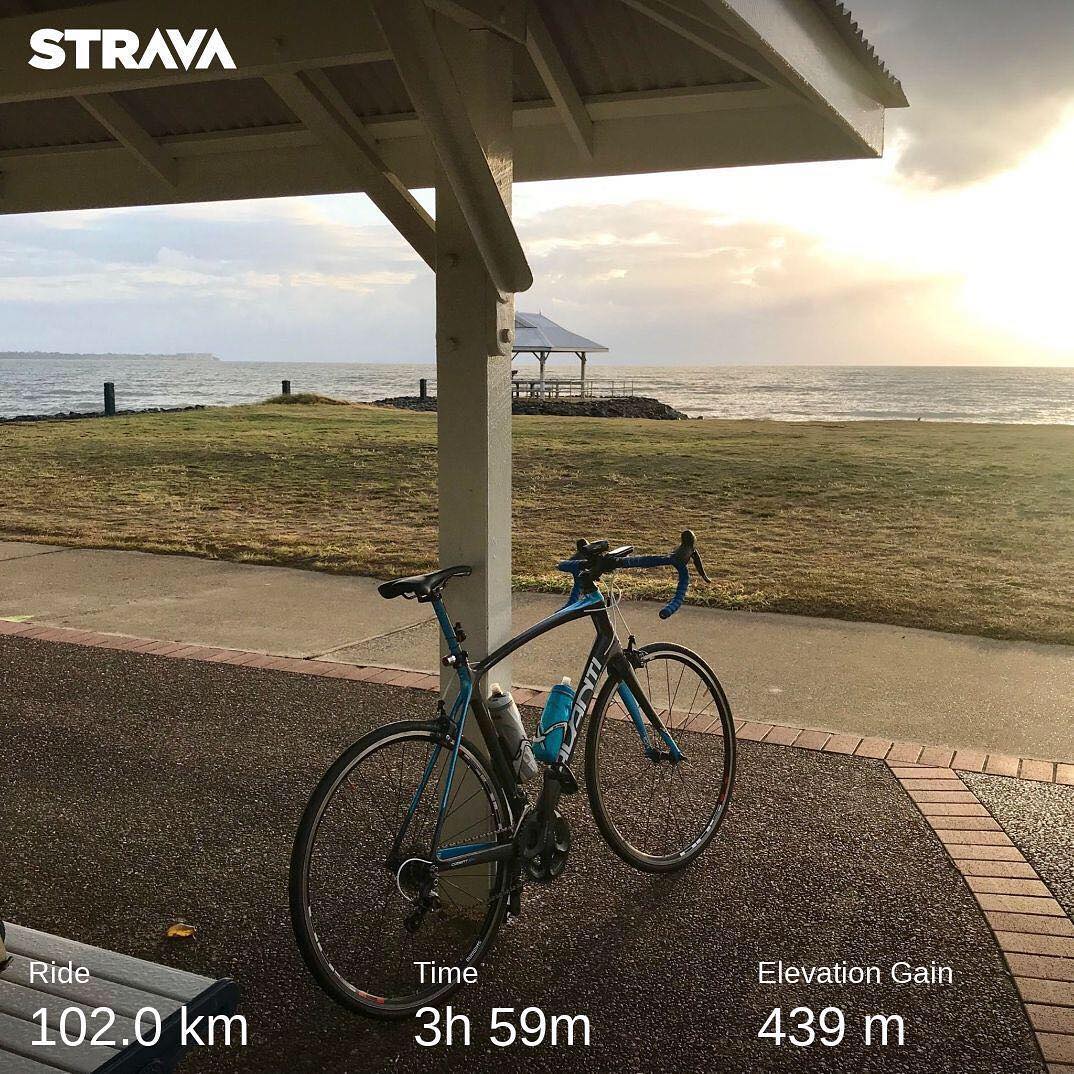 Road bike under a shelter by the ocean at sunset, with a grassy shore, small gazebo, and Strava ride stats overlaid.