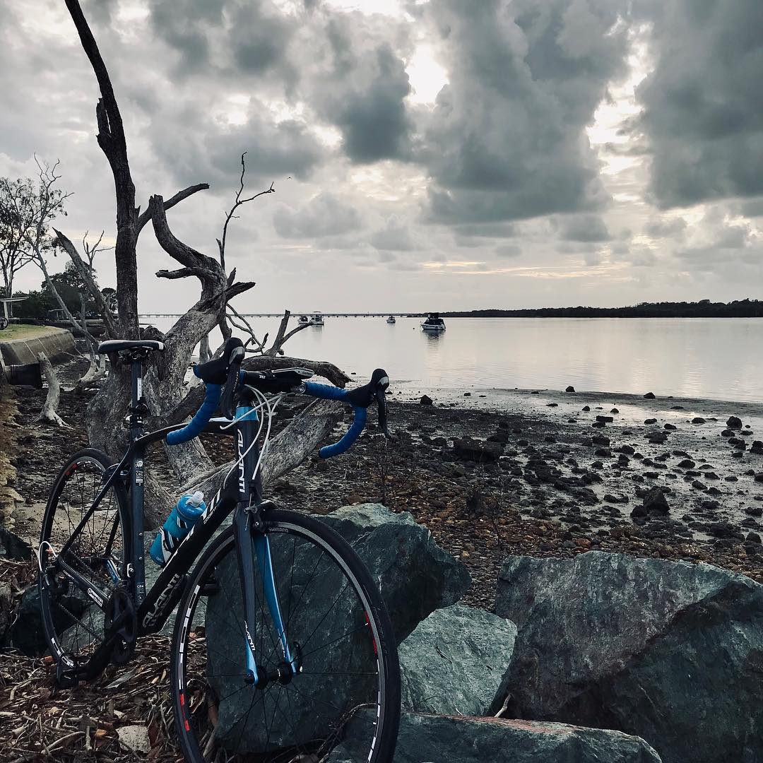 Road bike beside driftwood and rocks on a quiet shoreline under dark cloudy skies, with small boats on calm water in the distance.