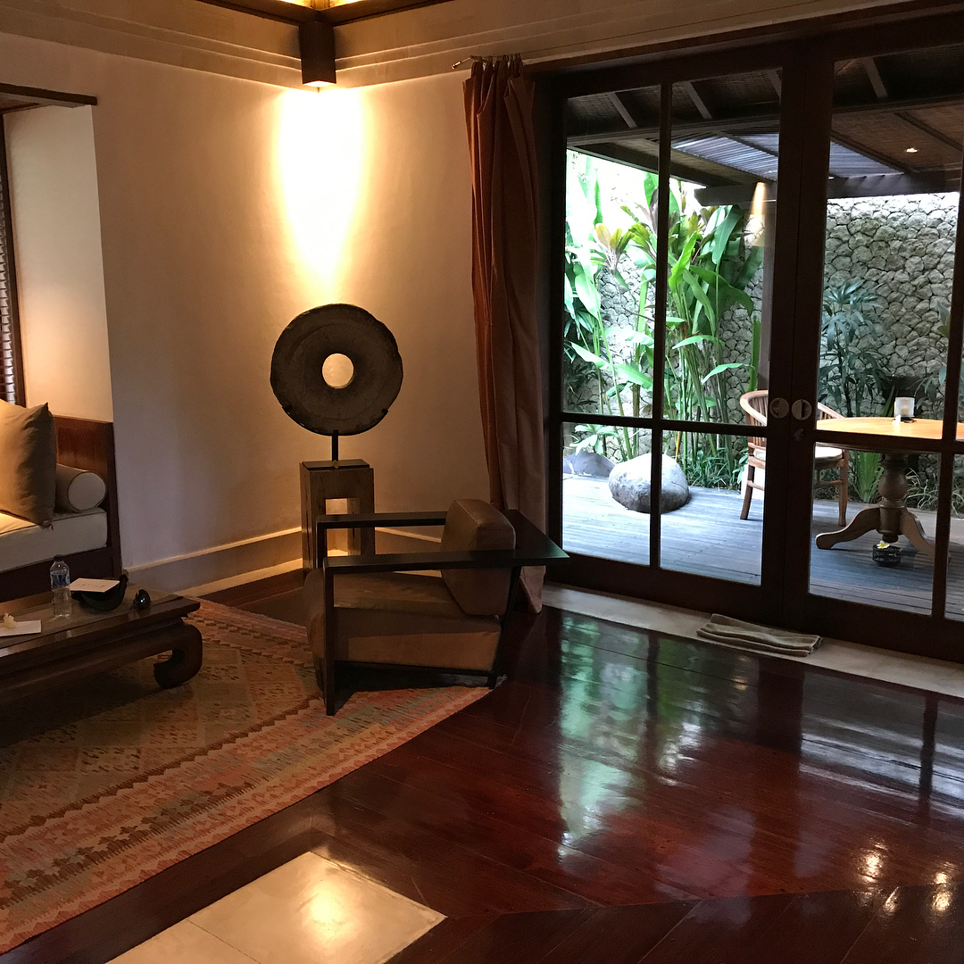 Warm living room with polished wood floors, a chair and sofa, and sliding glass doors opening to a small patio with plants and a table.