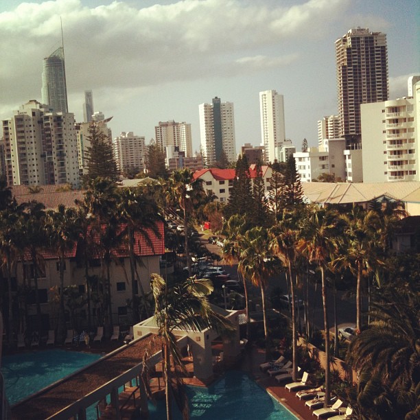 City skyline with tall apartment towers, palm trees, and a hotel pool area under cloudy skies.