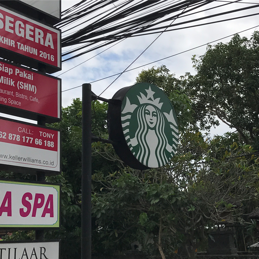 Starbucks round sign on a black pole beside red business signs, with trees and many overhead utility wires against a cloudy sky.
