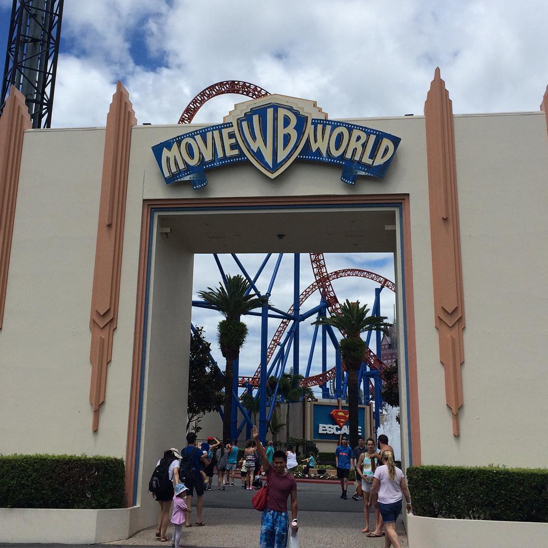Entrance arch with Movie World sign, people walking below, and a looping red roller coaster with blue supports behind it.