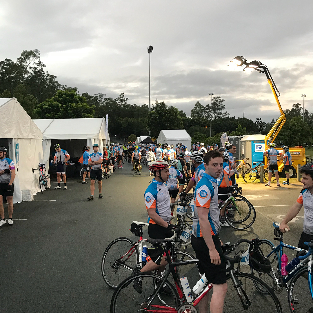 Cyclists in matching jerseys gather with road bikes near white tents and portable lights under a cloudy sky.
