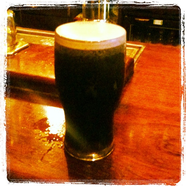 A pint glass of dark beer with a thick foamy head on a wooden bar top.
