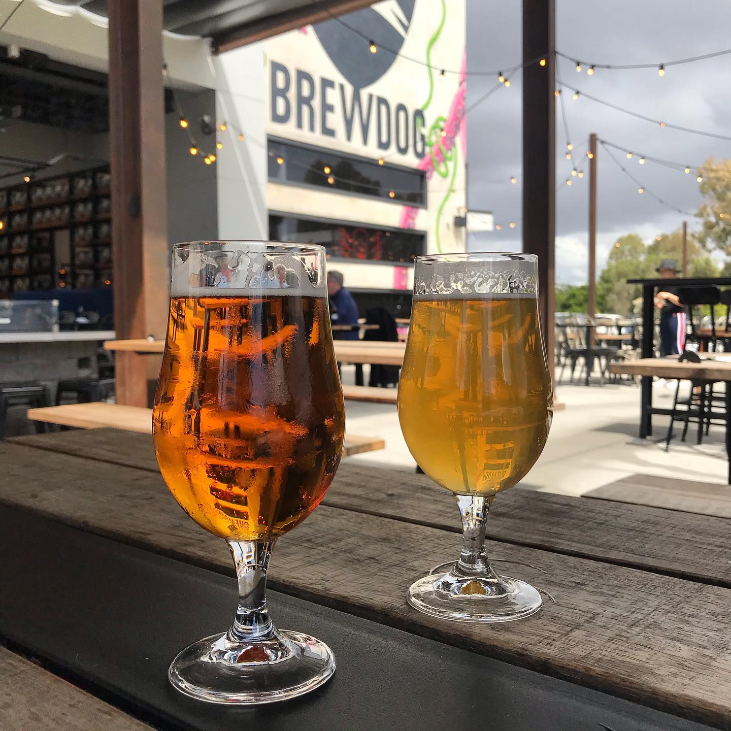 Two glasses of beer on a wooden table at an outdoor patio, with string lights and a BrewDog building in the background.