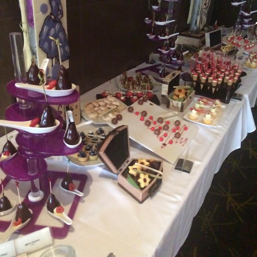 Dessert buffet on a long white table with tiered purple stands, chocolate-dipped treats, fruit cups, pastries, and mini cakes.