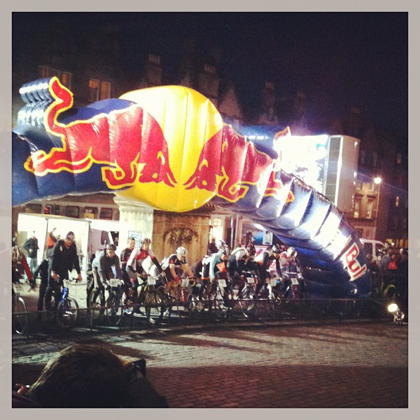 Cyclists line up at night beneath an illuminated Red Bull inflatable arch in a town square.