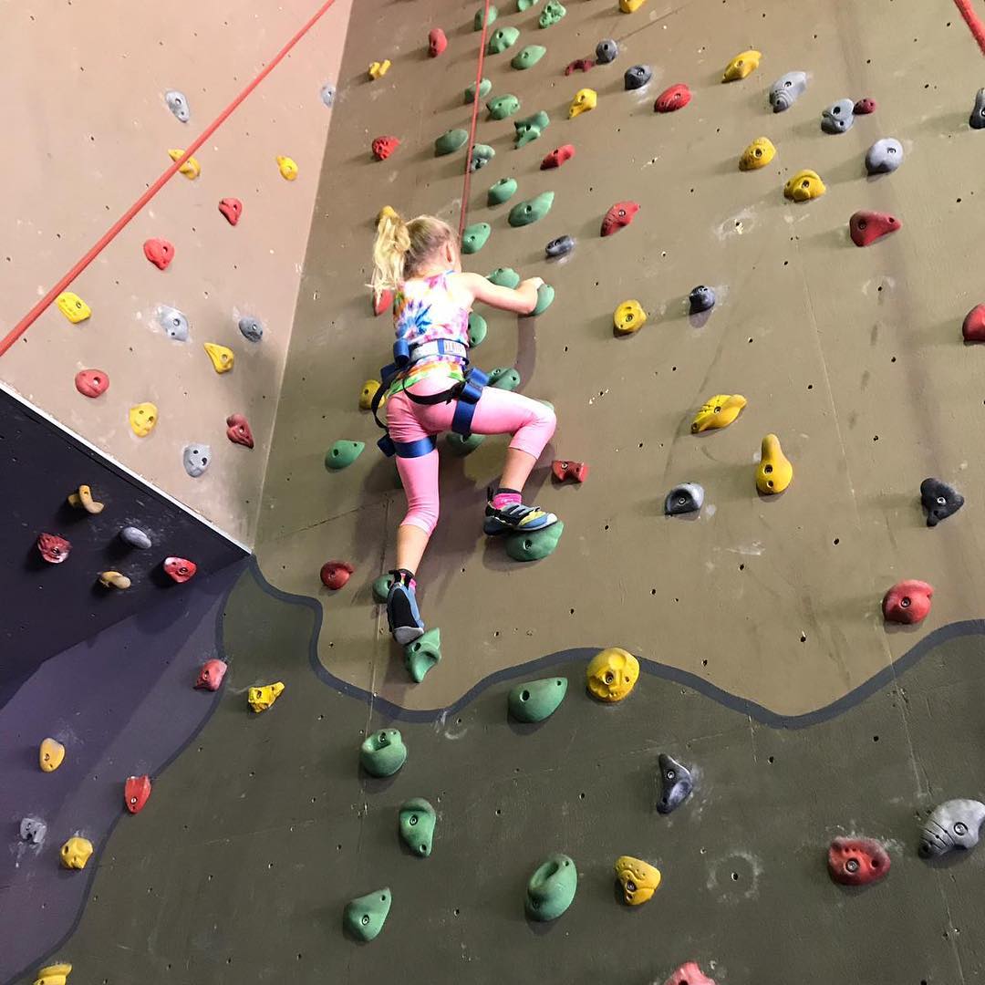 Child in a harness climbs an indoor rock wall with colorful handholds.