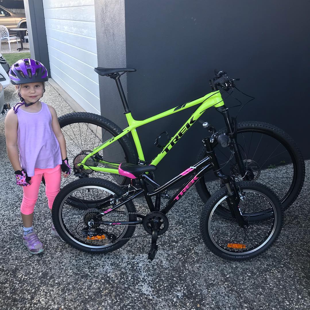 Young girl wearing a purple helmet stands beside a small black and pink bike in front of a larger bright green bike.