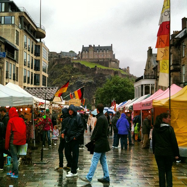 Rainy street market with colorful tents and shoppers, with a stone castle on a hill in the background.