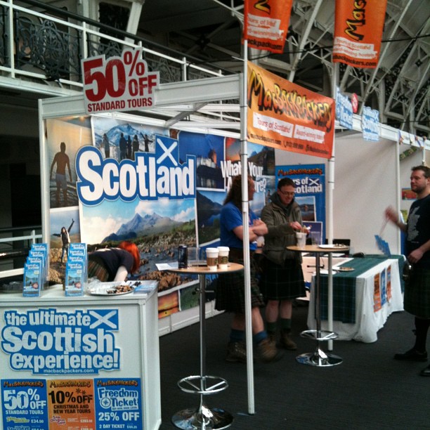 Scotland tourism booth at an indoor expo with large signs, discount offers, and people standing by tall tables.