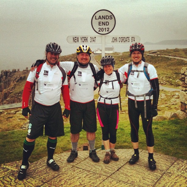 Four cyclists in matching jerseys stand smiling beneath the Land’s End sign on a rocky coastal path.