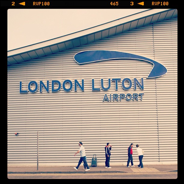 Exterior wall with large "LONDON LUTON AIRPORT" sign and four people walking along the sidewalk below.