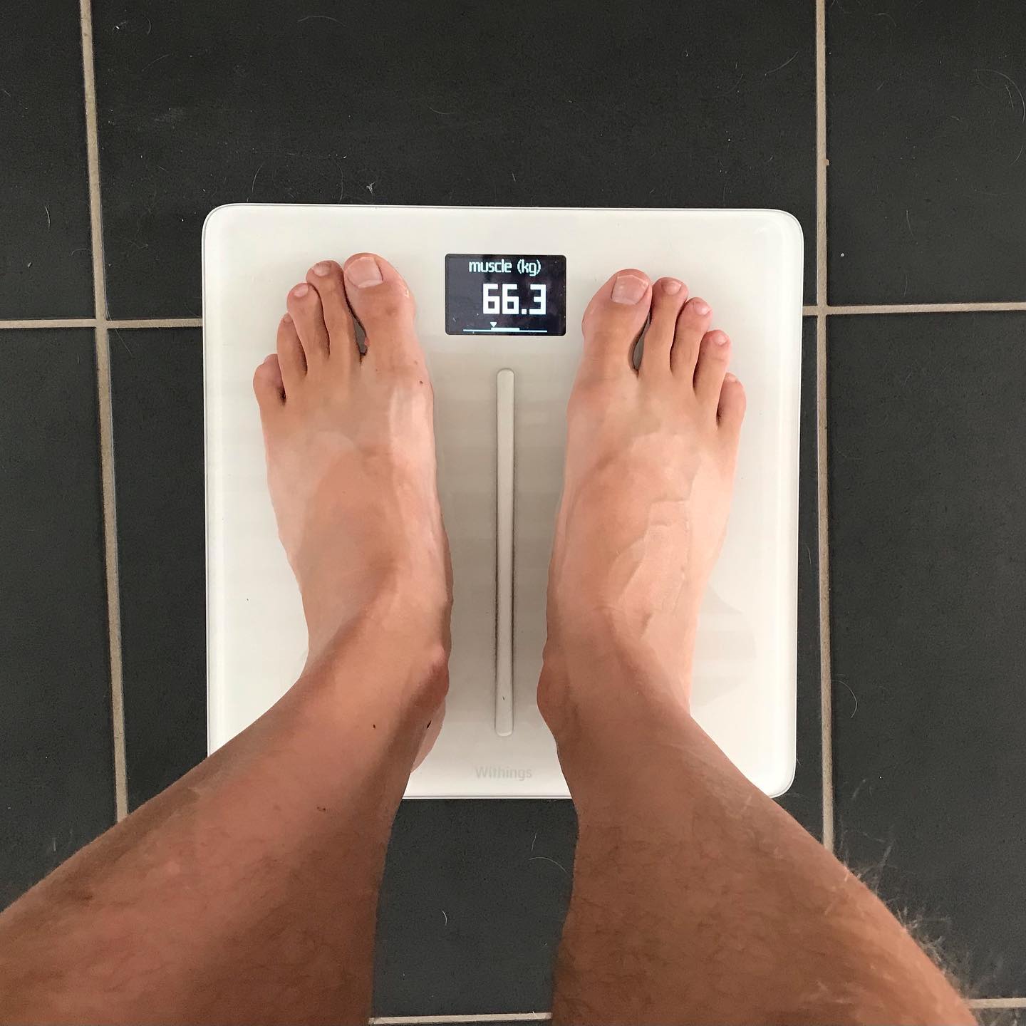 Bare feet standing on a white digital scale on dark tile floor, with the display showing muscle 66.3 kg.