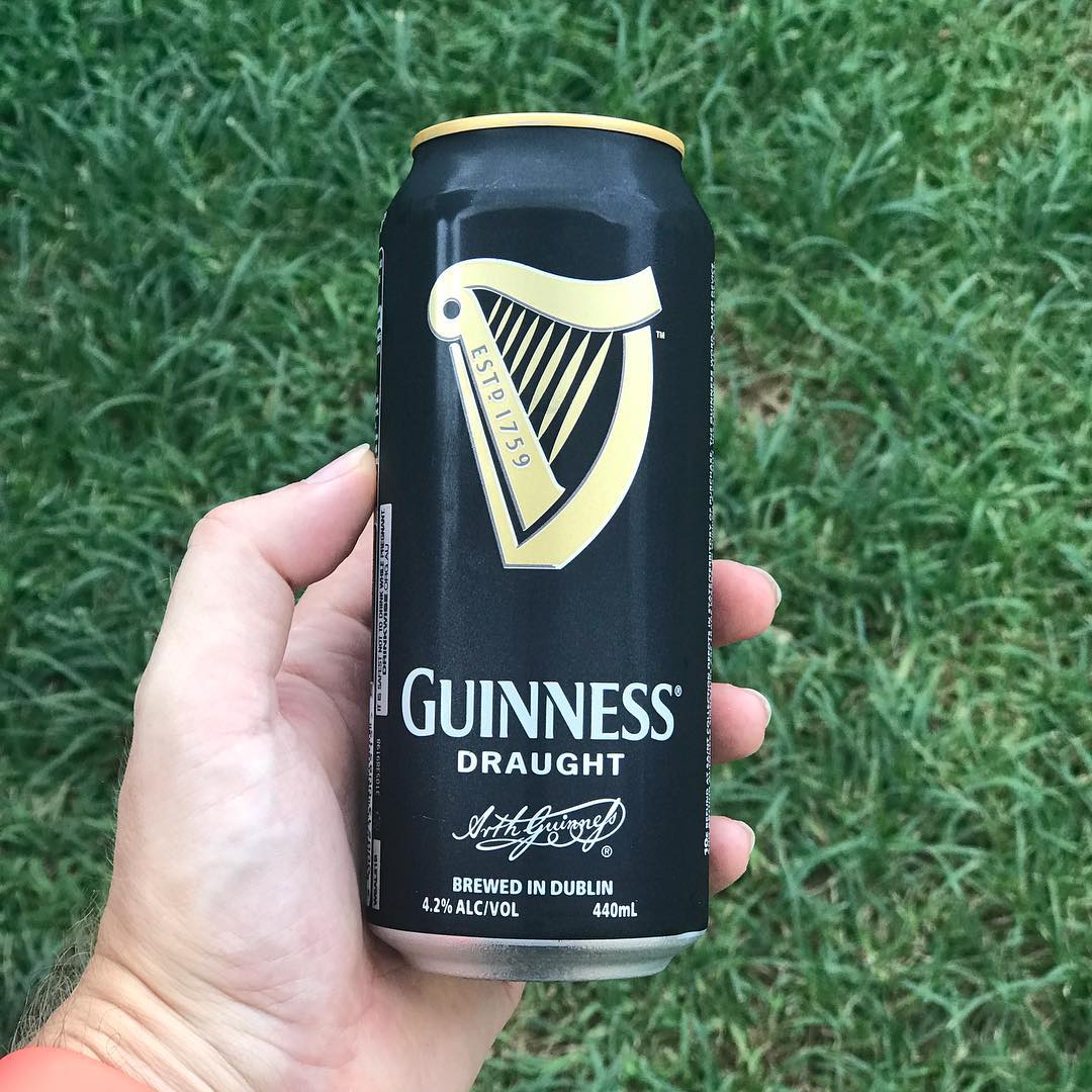 Hand holding a black Guinness Draught can with gold harp logo against a grassy background.