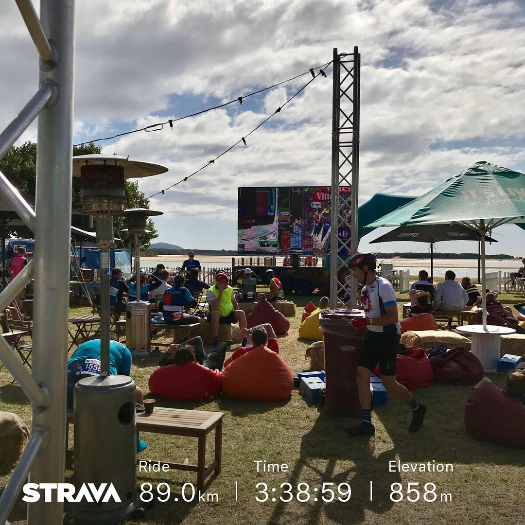 People relaxing on beanbags by the water under umbrellas, with cyclists, patio heaters, and a large screen; Strava stats overlay at bottom.