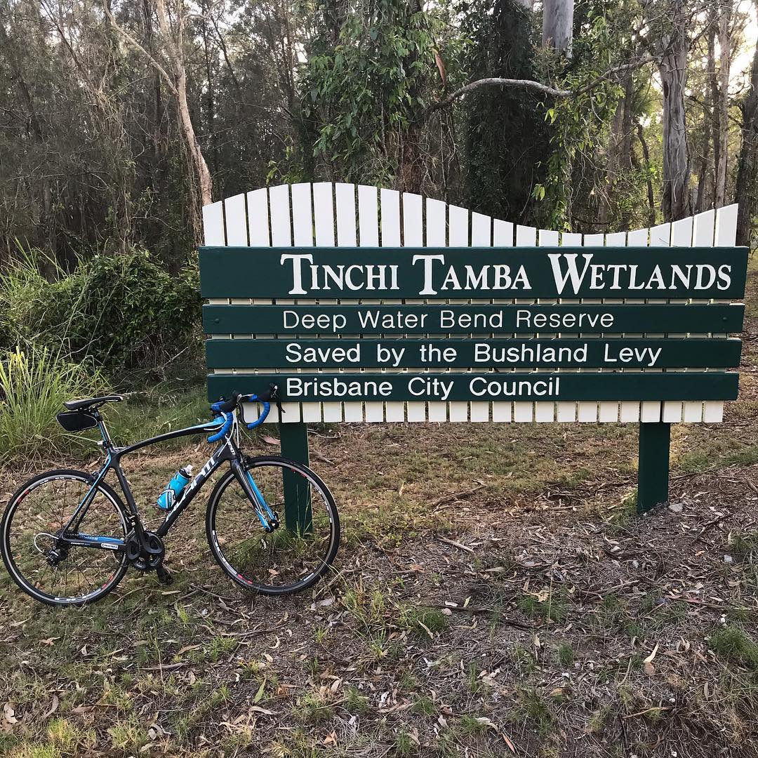 Road bike leaning against a sign reading “Tinchi Tamba Wetlands” in a wooded area.