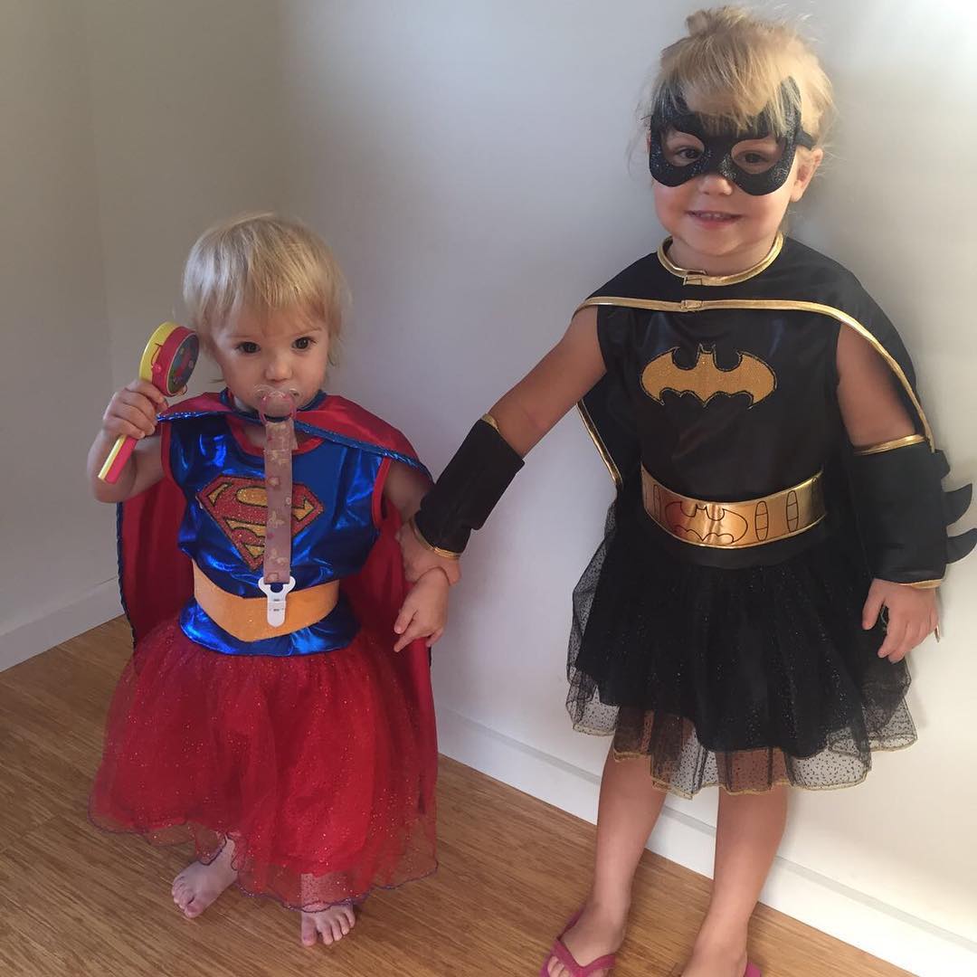 Two toddlers holding hands in Supergirl and Batgirl costumes, with one wearing a pacifier clip and holding a toy rattle.