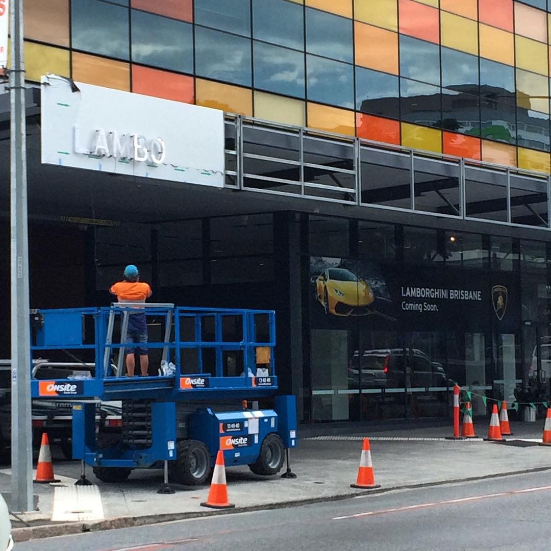 Worker on a blue lift removing letters from a white sign outside a building with colorful glass panels and orange traffic cones.