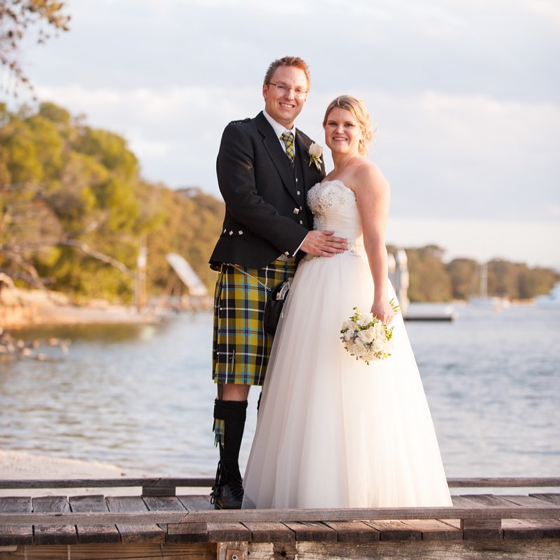 Bride in a white gown holding a bouquet stands with a smiling man in a kilt on a wooden dock beside calm water.