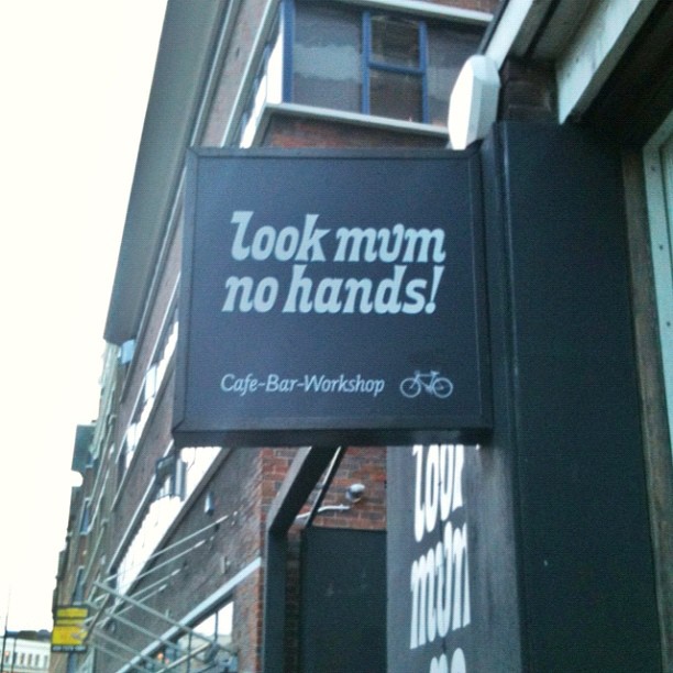 Black hanging storefront sign reading “look mum no hands!” with smaller text “Cafe-Bar-Workshop” and a bicycle icon.