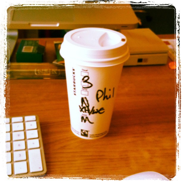 Starbucks paper coffee cup with “Phil” written on it, sitting on a wooden desk beside a keyboard.
