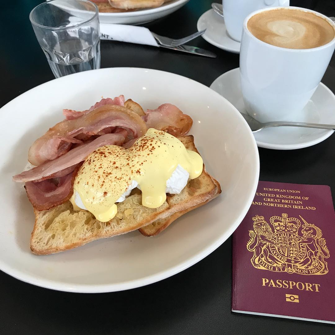 Plate of eggs Benedict with bacon and toast beside a cappuccino, water glass, and a UK passport on a black table.