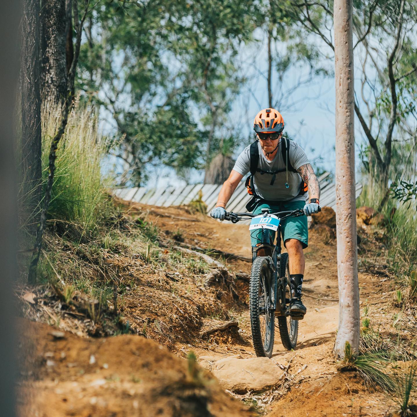 Cyclist wearing an orange helmet rides a mountain bike down a narrow dirt trail between trees.