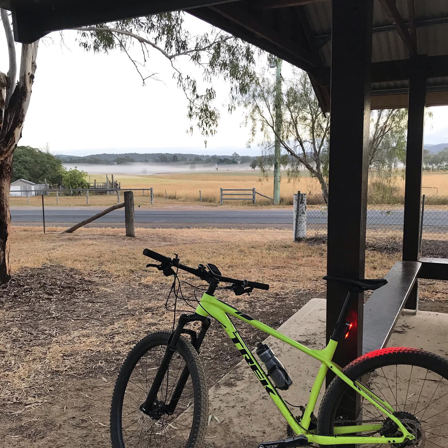 Bright green mountain bike under a shelter beside a road, with dry fields, trees, and low mist in the distance.