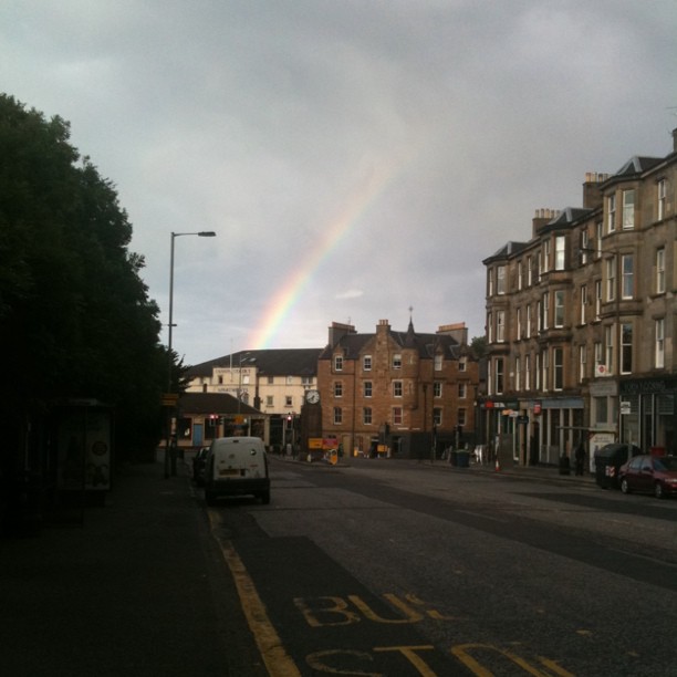 Rainbow over a quiet city street lined with stone buildings, parked cars, and dark trees under a cloudy sky.
