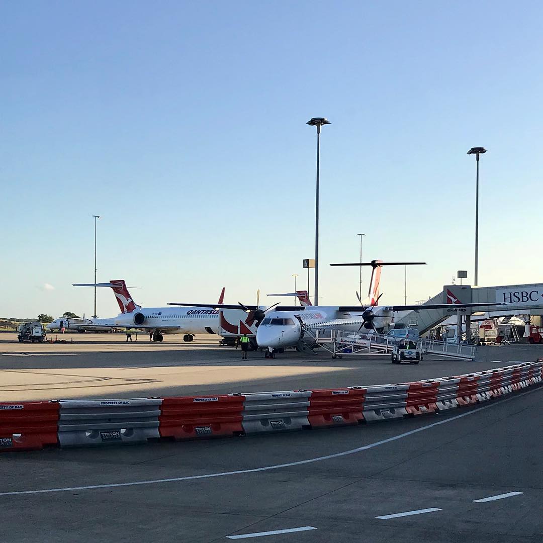 Qantas planes parked on an airport tarmac beside a jet bridge, with red and white barriers in the foreground.