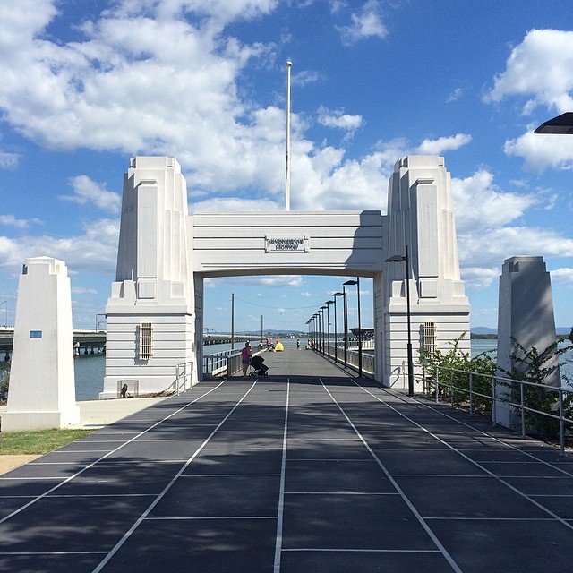 White art deco gateway at the end of a tiled waterfront walkway under a blue sky with scattered clouds.