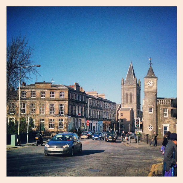 Cars and pedestrians on a city street lined with stone buildings, including a church tower and a clock tower under a clear blue sky.