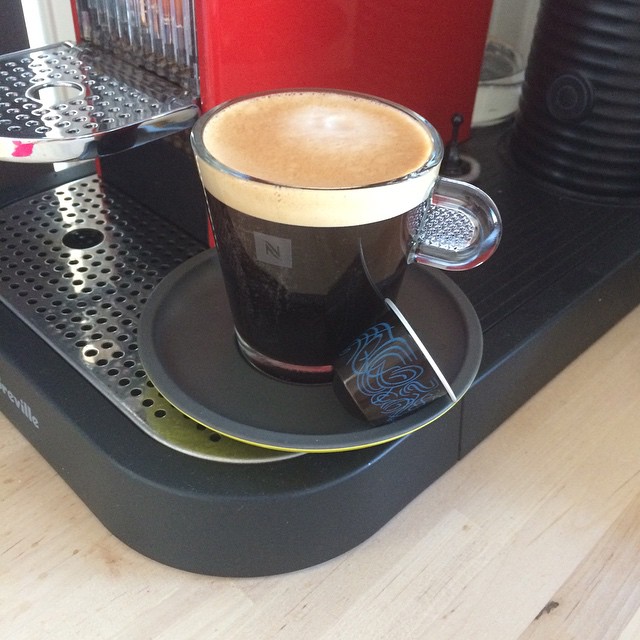 Glass cup of black coffee with crema on a saucer and wrapped chocolate on a coffee machine drip tray.