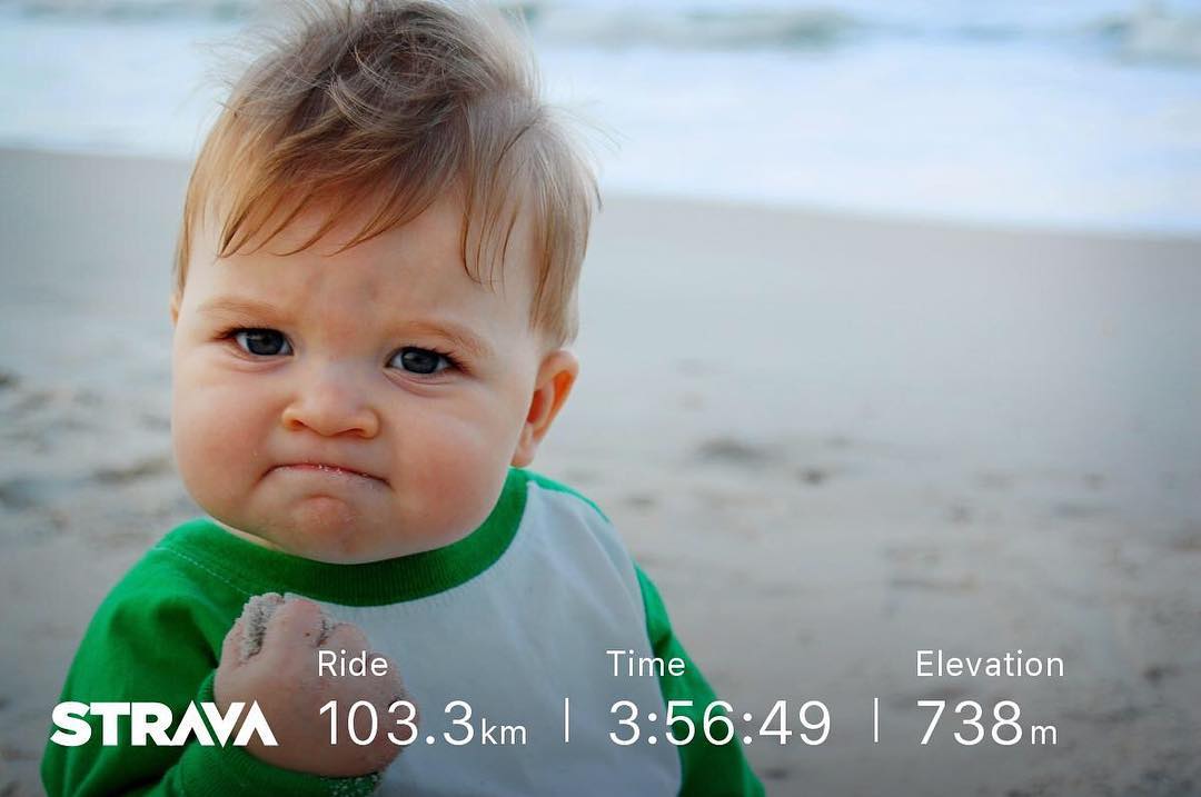 Baby with a clenched fist on a beach, overlaid with Strava stats: ride 103.3 km, time 3:56:49, elevation 738 m.
