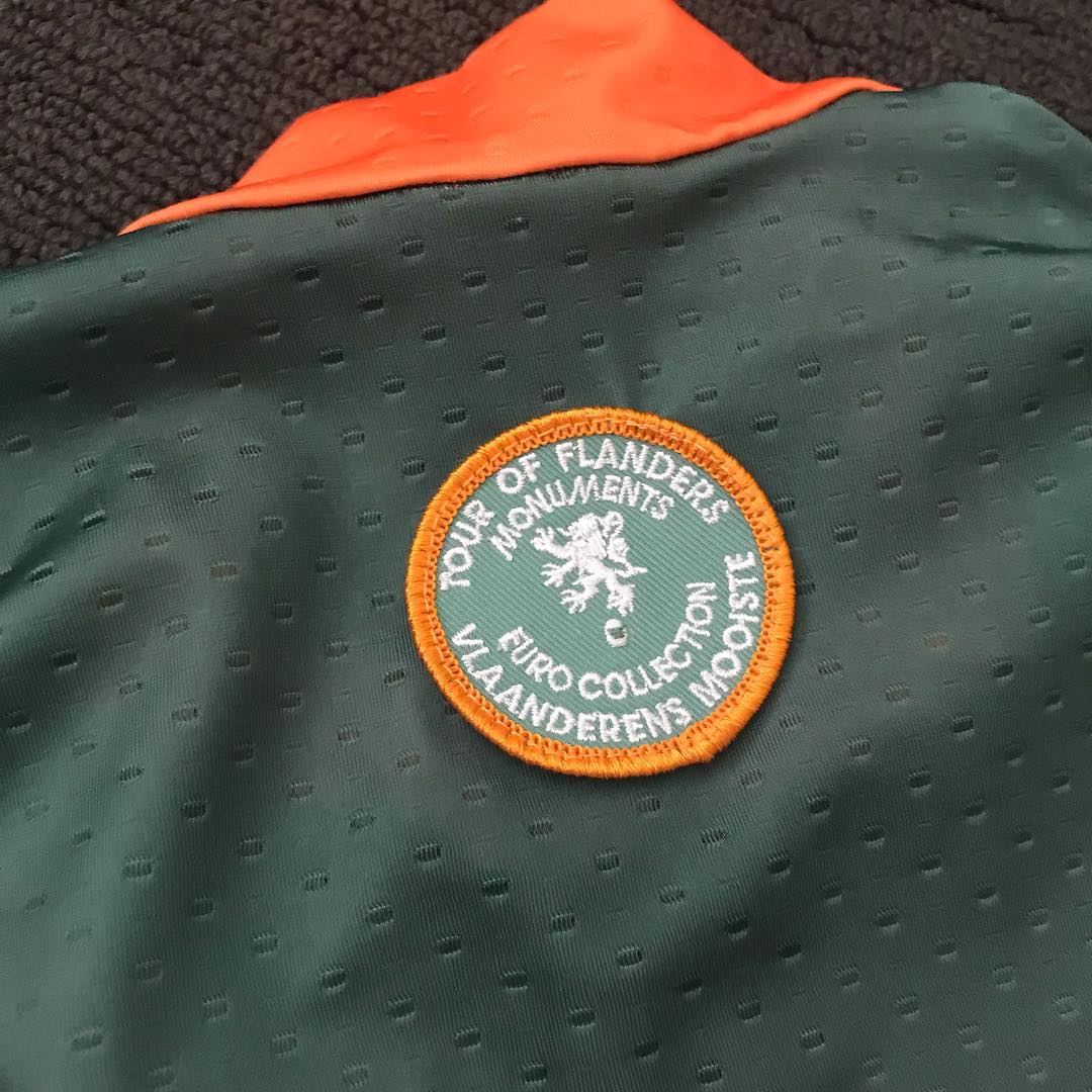 Green perforated fabric with an orange edge and a circular embroidered patch reading “Tour of Flanders Monument” and “Vlaanderen Mooiste.”