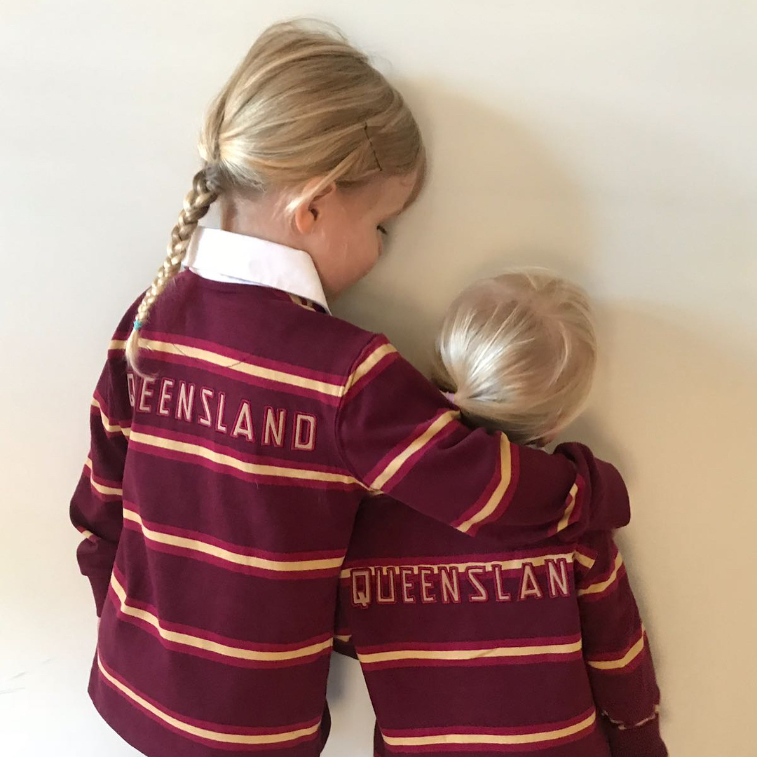 Two blonde children in matching maroon striped shirts with "Queensland" on the back stand side by side, with the older child hugging the younger.