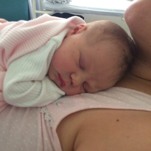 Sleeping newborn in a white outfit and pink blanket resting on an adult’s chest.