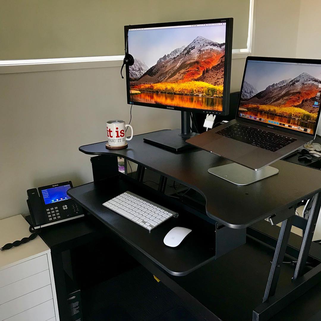 Black standing desk setup with external monitor, MacBook on a stand, keyboard, mouse, office phone, headset, and a mug.