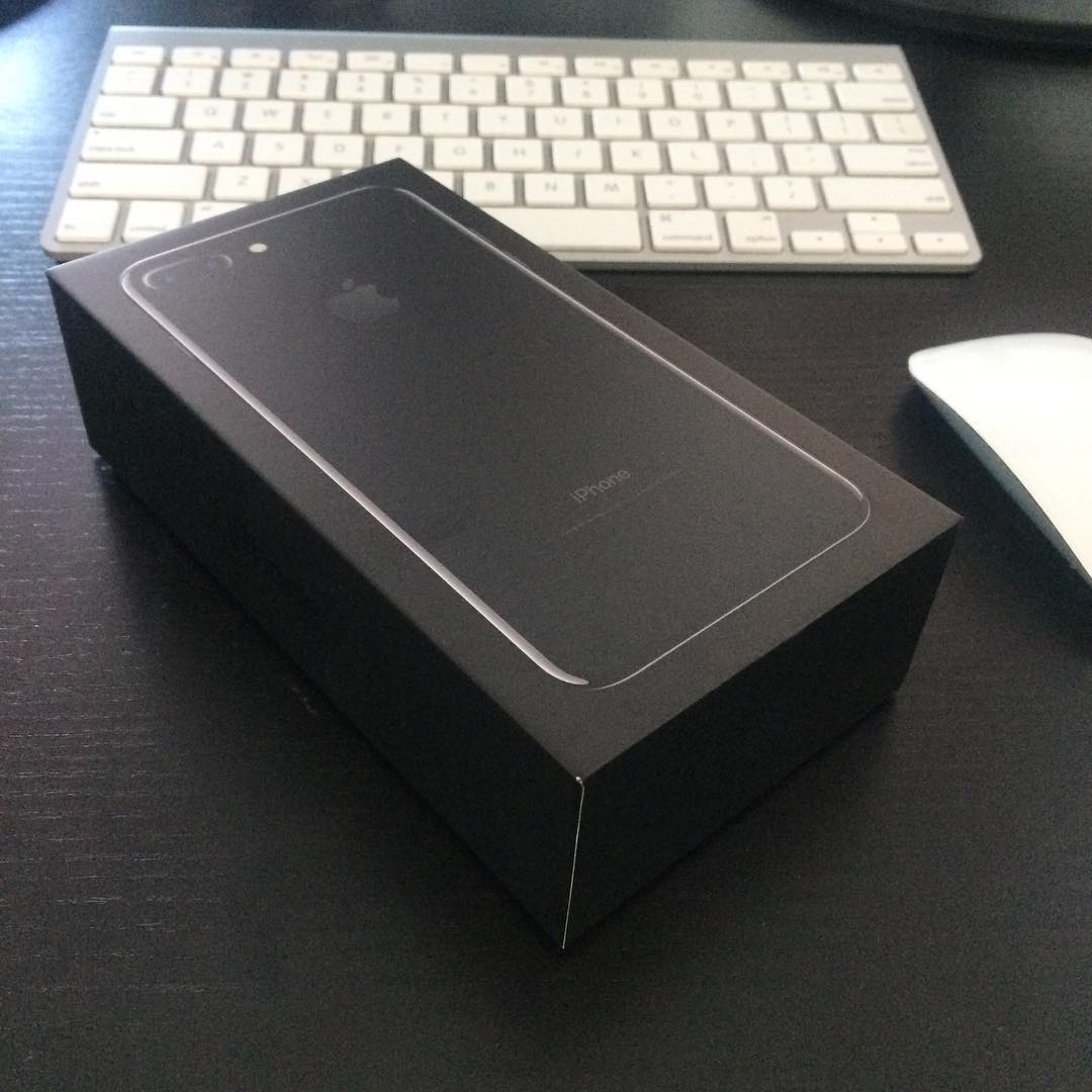 Black iPhone box on a dark desk with a white keyboard behind it and part of a white mouse beside it.